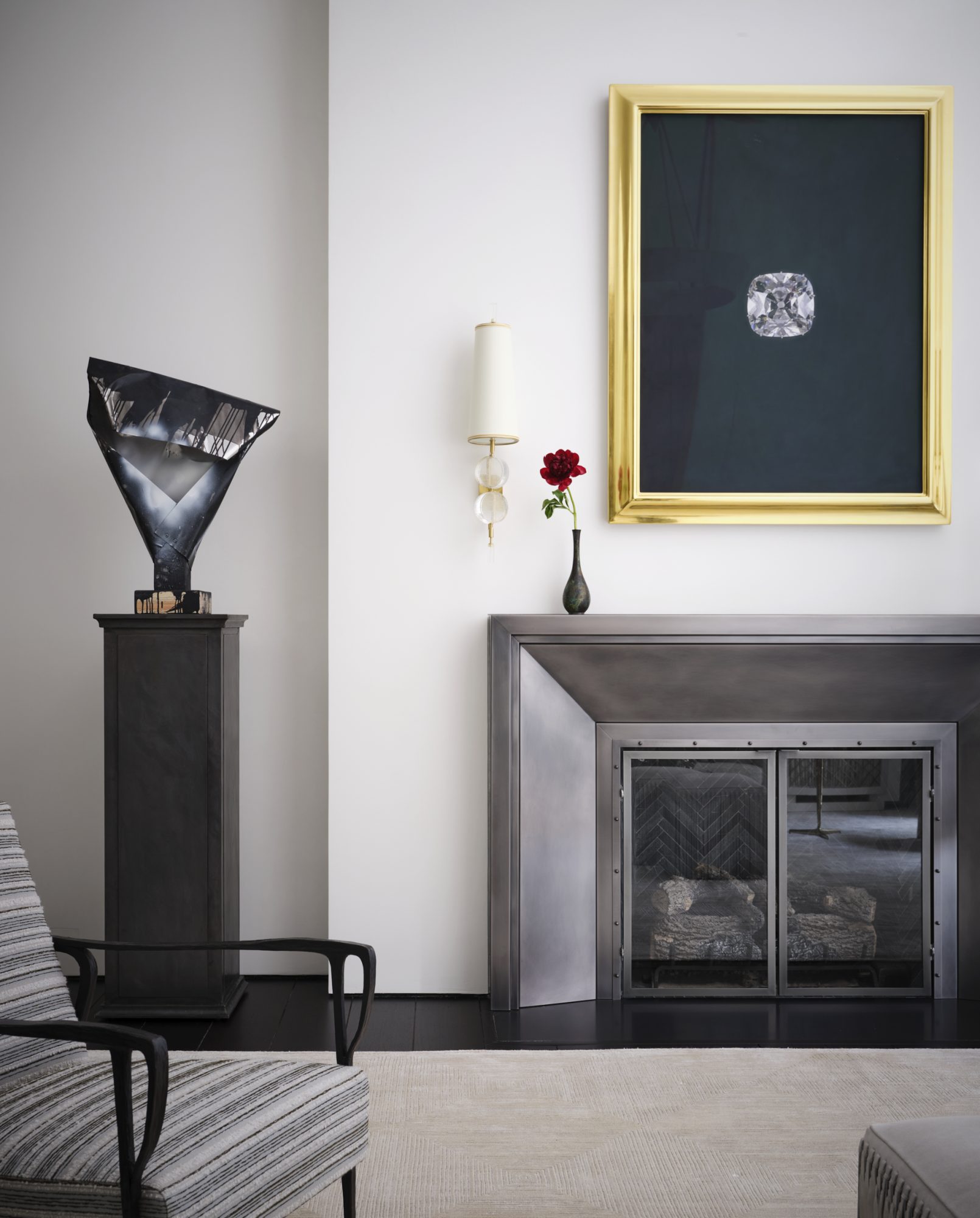 Living room setting with striped modern chair, steel art on a pedestal, a steel fireplace surround with a flower vase on the mantel, white shade sconce on the wall and gilded frame artwork showing an enlarged single gemstone on the wall.