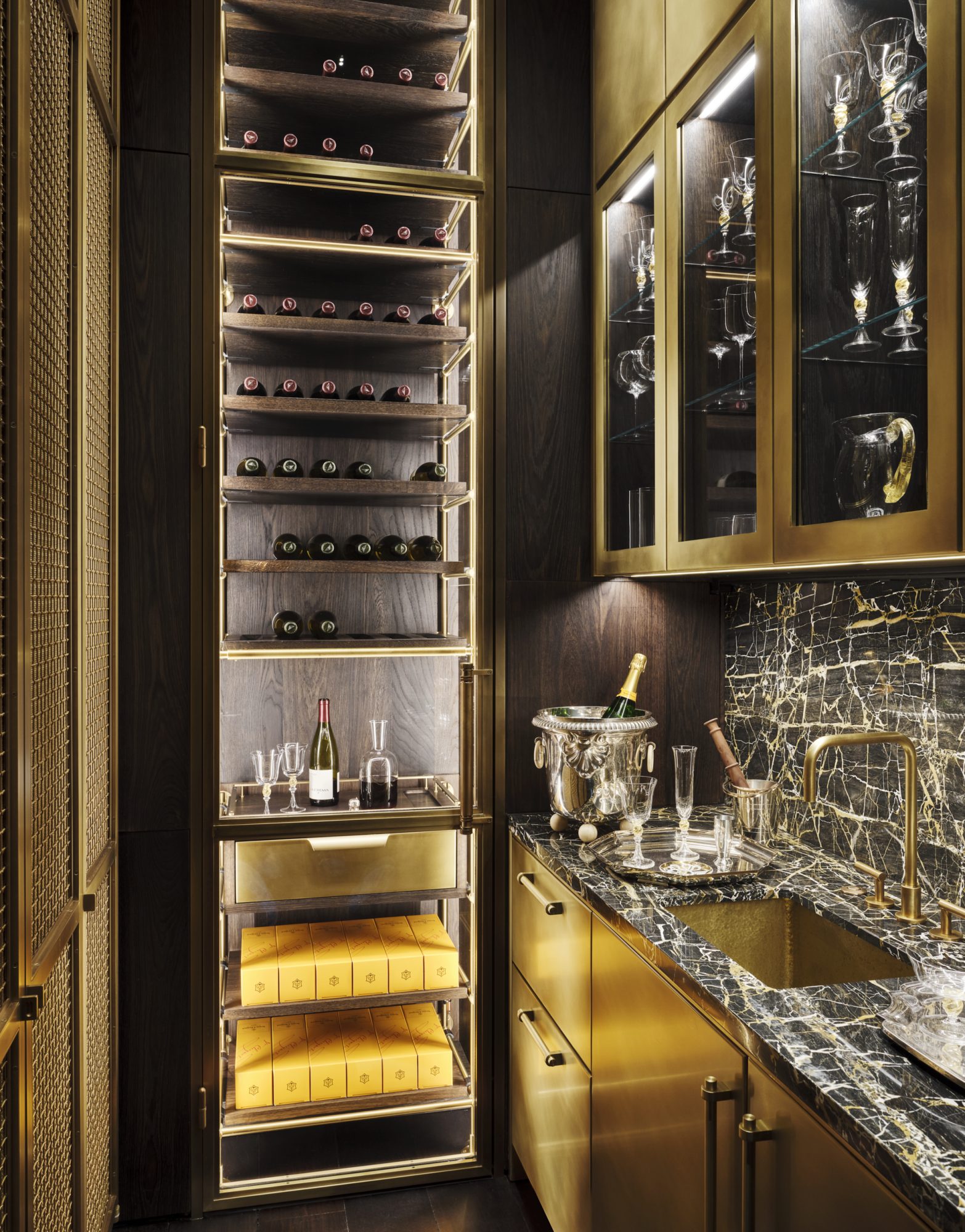 Amuneal's bronze butler's pantry in a narrow space of a Manhattan town house. Open Wine Shelving in a back lit display with carved oak wine shelves and a bronze sommelier station work surface and drawer. Stands floor to ceiling behind glass with a bronze door frame. Bronze clad bar cabinetry on the right shows upper glass and brass cabinets alongside black stone with white veining.