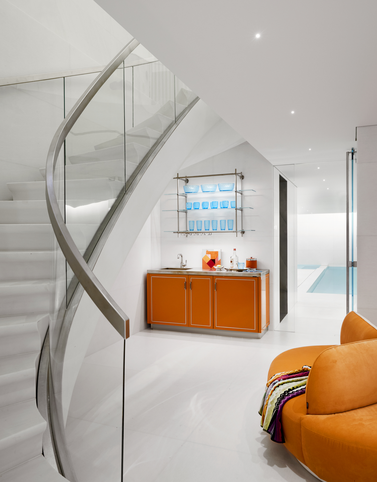 Curved stairs lead down to a basement pool house with an orange round couch and an hermes orange lacquer bar with champagne brass inlay, counter and brass and glass shelving above.